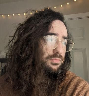 A man with long, dark brown hair and a full beard stares off into the distance through clear rimmed glasses. His eyes are obscured by the light reflecting in the lenses.