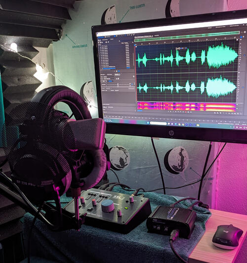 A recording setup. Headphones sit on a microphone stand that holds a Neumann TLM103 microphone. A small audio interface sits on a raised desk. Adobe Audition is on a monitor showing a recently recorded waveform.